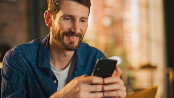 一个英俊男子坐在沙发上的肖像，使用智能手机在时尚的阁楼公寓或咖啡馆。创意男性微笑，查看社交媒体和输入信息。从大窗看城市。图片下载