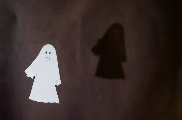 一个白纸鬼和他的影子在灰色的墙上。用剪刀从一张纸上剪下的万圣节用具。在空中飞翔的幽灵，画着嘴和眼睛的剪影。图片下载