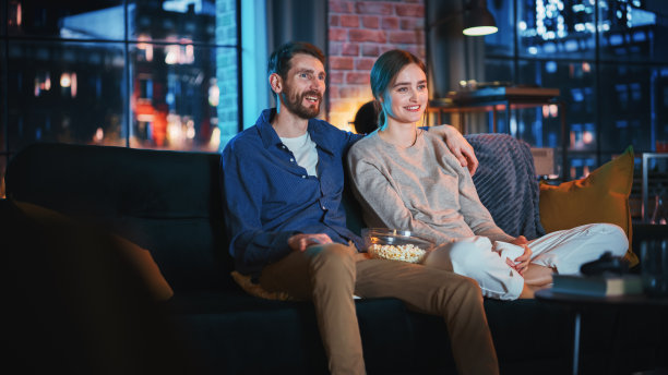 美丽夫妇的肖像花时间在家里，坐在沙发上，拥抱和看精彩的电视节目在他们的时尚阁楼公寓。男人和女人流媒体喜剧电影和有好的时间。图片下载