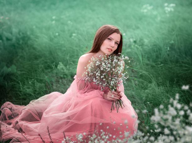一个年轻的女孩走在一片白花的田野里。这个女孩手里拿着一束花。这个女孩穿着一件粉红色的长连衣裙。图片下载