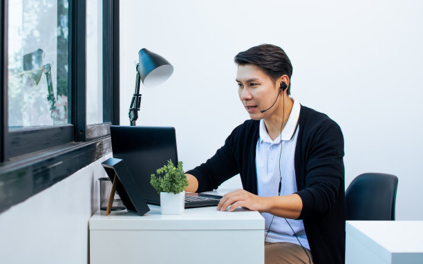 穿着休闲衬衫，戴着耳机，在家用笔记本上网学习或教学，面带积极快乐的微笑的亚洲帅哥。新常态、新科技理念。图片下载