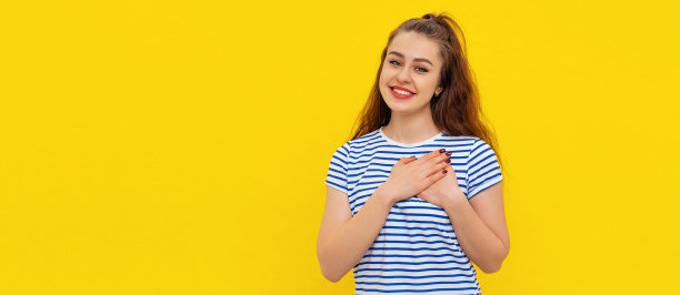 肖像和平，亲爱的女孩手握在胸前接近心脏，微笑感激，显示赞赏和爱，站在条纹t恤衫在黄色背景与复制自由空间的文本图片下载