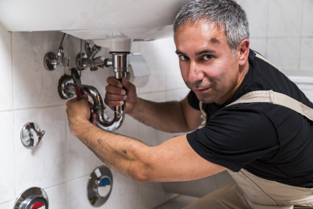 一名专业男水管工的肖像，他穿着工装裤和黑色t恤，脸很脏，正在修理浴室水槽下的水龙头图片下载