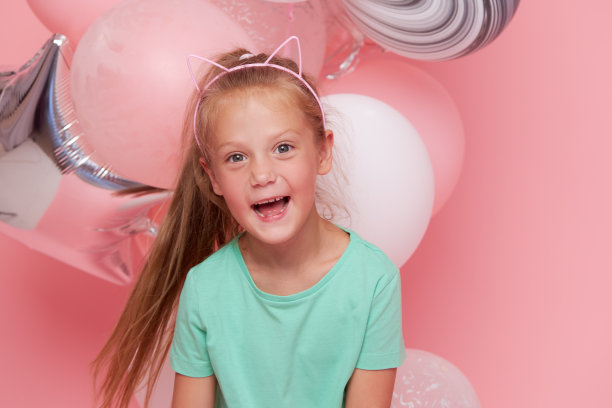 欢快的小女孩穿着优雅的薄纱裙靠近气球。生日礼物。有趣的脸庆祝生日聚会在粉红色的背景图片下载