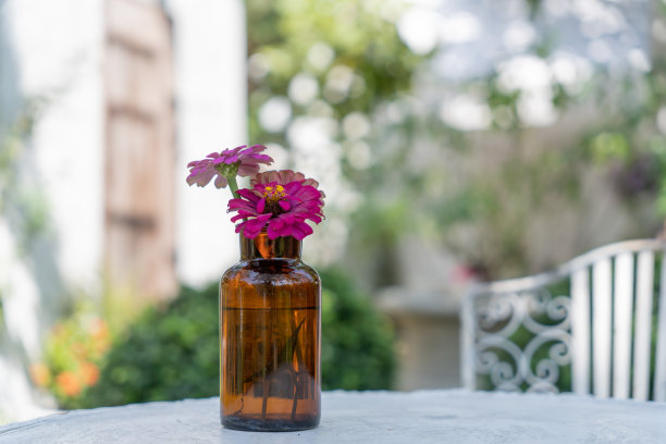 小瓶鲜花放在花园的白色桌子上。春天静悄悄地开着花。花朵在棕色玻璃与自然背景。图片下载