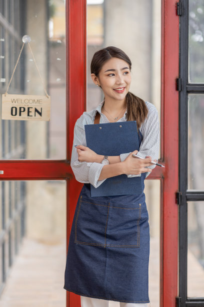 照片中微笑的年轻咖啡馆老板亚洲女人站在门口，双手交叉。打开玻璃门上的招牌。图片下载