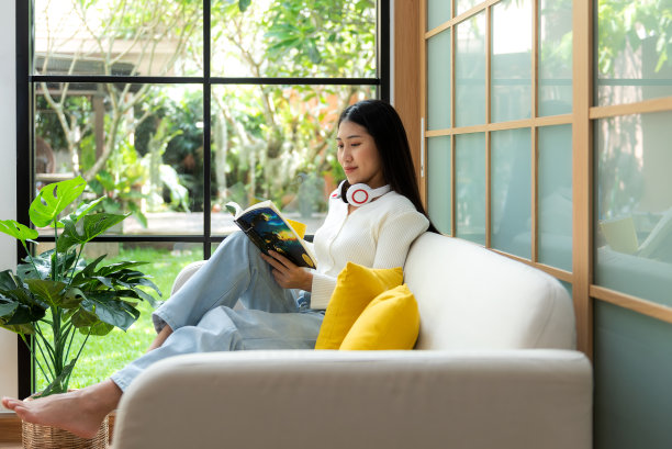 快乐的年轻女子在家里的沙发上看书。生活方式自由，在客厅喝咖啡放松和冷静，自然花园背景。生活方式的概念图片下载