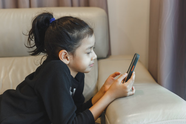 小女孩躺下玩看智能手机在线学习。小女孩在线聊天学习。屏幕上的小女孩，社交媒体成瘾。图片下载