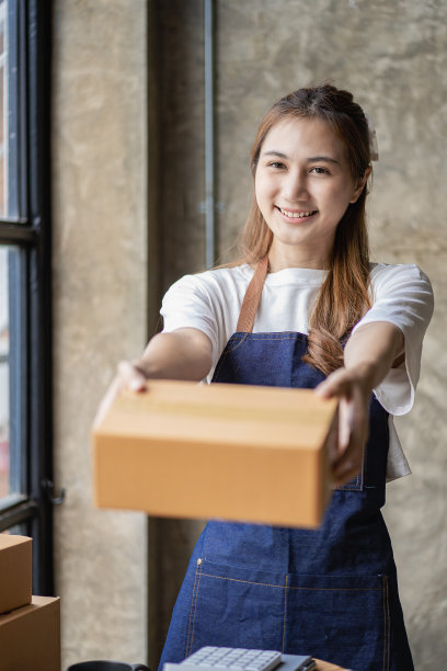 一名亚洲妇女，在家里摆弄着盒子和笔记本电脑。她开办了一家中小企业，在网上用包裹递送推销自己的想法。垂直图图片下载