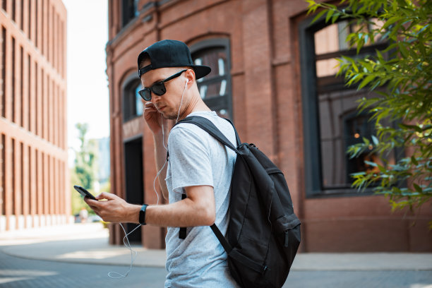 年轻人是一个时髦的家伙，戴着墨镜和黑色的帽子在城市里走，手里拿着智能手机，听着音乐耳机。T图片下载