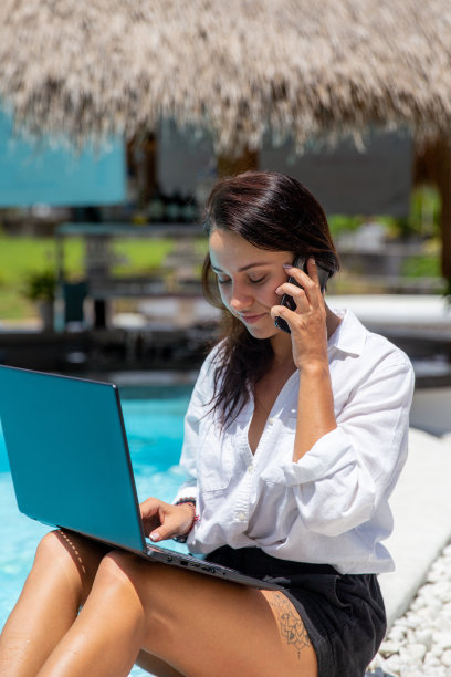 年轻的白人女性在靠近蓝色游泳池的地方远程工作，同时使用笔记本电脑和手机。图片下载