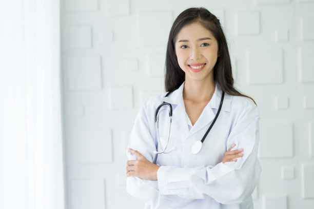 快乐的亚洲护士穿着带听诊器的制服，双臂交叉站在医院里。图片下载