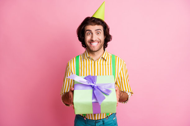 年轻兴奋的阿拉伯男子的照片快乐的微笑持有礼物礼物戴派对帽子孤立在粉红色的背景图片下载