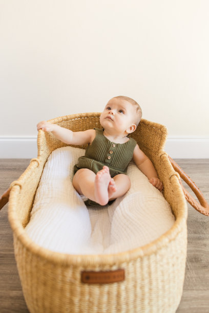 一个27周大的小男孩穿着橄榄绿针织连体裤，坐在一个舒适的棉毯上，在一个中性的现代家庭的海草摩西篮子里图片下载