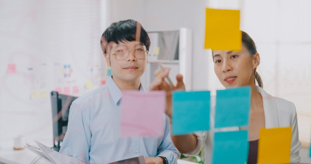 年轻的亚洲商人和女商人团队合作，正装衬衫站在透明的白板头脑风暴会议和讨论的商业计划在小的工作场所。图片下载