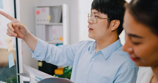 特写年轻的亚洲商人和女商人团队手持电子平板，站在透明的白板前进行头脑风暴会议，并在工作场所撰写商业计划。图片下载