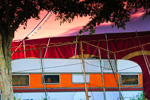 橙色条纹的大篷车停在围栏后面，旁边是马戏团的红色篷布。图片下载