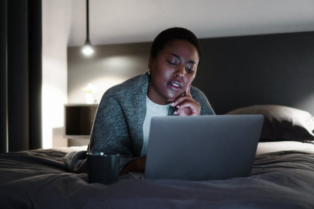 微笑的黑人女性，端着一杯咖啡，躺在床上，看着笔记本电脑的屏幕图片下载