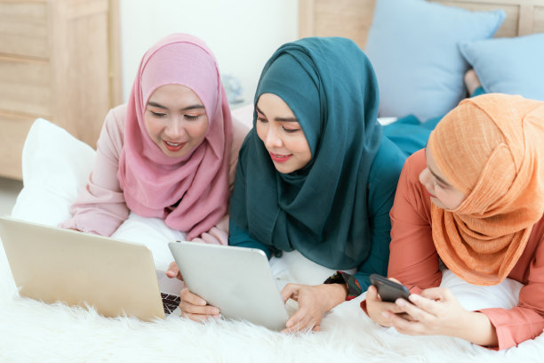 一群穆斯林妇女躺在床上，用笔记本电脑愉快地聊天。图片下载