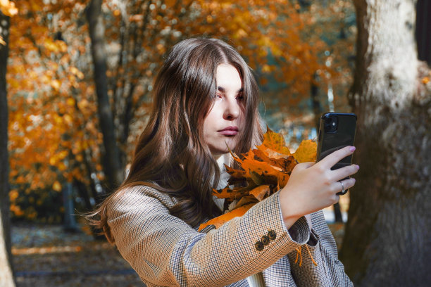可爱的年轻严肃的女人在秋天的森林在电话上自拍。图片下载