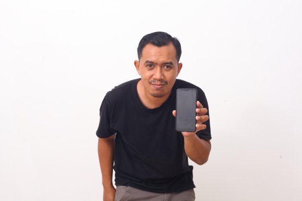 满脸胡须、身穿黑色t恤的亚洲男子满意地站在手机前，微笑着向相机推荐小工具或移动应用程序。室内工作室拍摄孤立的白色背景图片下载