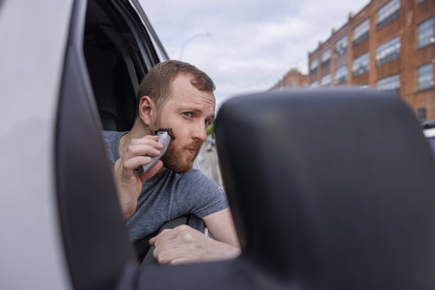 一名司机在停在街上的卡车里用后视镜和便携式修剪器刮胡子。图片下载