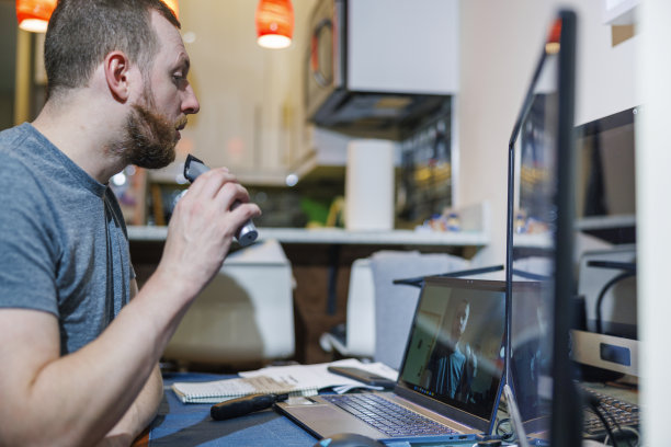 一名白种白人男子是电脑爱好者，他在公寓的客厅里用无线修剪器刮胡子，把笔记本电脑的摄像头和显示器当作镜子。图片下载