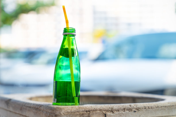 在停车场前的街道上，水泥垃圾桶的边缘上立着一个装着未喝完饮料的玻璃瓶和伸出来的吸管，近景，模糊的背景。图片下载