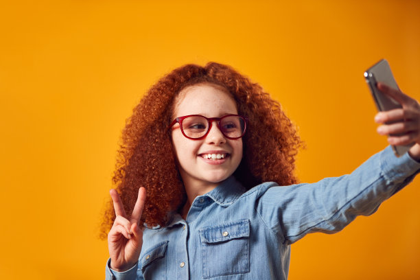 在黄色背景下，戴眼镜的女孩在手机上自拍的工作室肖像图片下载