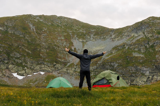 山上的帐篷旁有一个人举起双手。帐篷搭在旁边。被群山包围着。色彩鲜艳的旅游帐篷在大自然的群山中图片下载