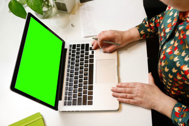 分散注意力的女人拿着笔，在办公室用电脑工作。笔记本电脑绿屏。一天工作的开始或结束。工作区。文书工作。工作的时候了。特写镜头的女商人。的焦点图片下载