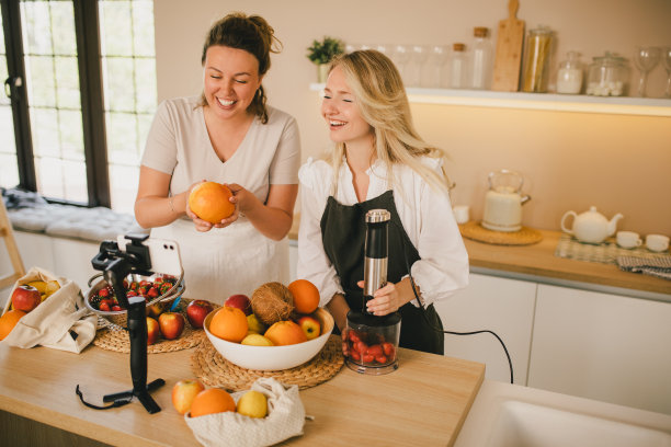 两位千禧一代的女博主在厨房用智能手机录制制作奶昔的视频。图片下载
