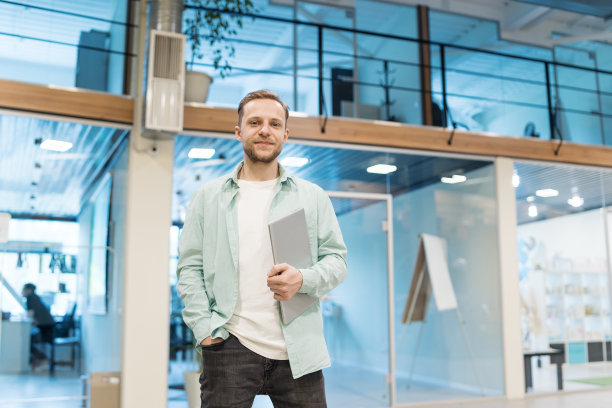 自信的男性企业家站在现代化的办公大厅里看着镜头。年轻人在不经意间拿着笔记本电脑，手插在口袋里图片下载