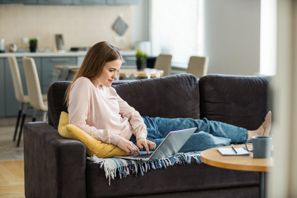 美丽的年轻女子在家里躺在沙发上用笔记本电脑工作或学习的侧面视图。女性躺在沙发上用电脑打字图片下载