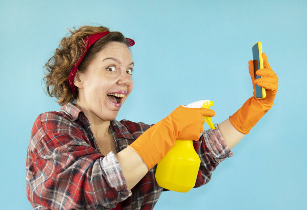 在蓝色的背景上，身穿格子衬衫的欢快的成年美女手里拿着喷枪和手机。美女的女人肖像图片下载