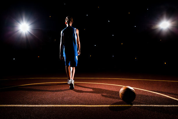 篮球。一个穿着蓝色运动服的少年从一个躺在运动场上的篮球旁边走了出来。背景中有两盏聚光灯。黑暗的背景。体育游戏的概念图片下载