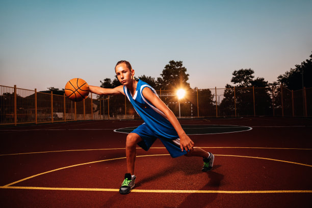 篮球。身穿蓝色运动服的少年正在自信地打篮球。背景是一片运动场地和暮色。体育游戏的概念图片下载