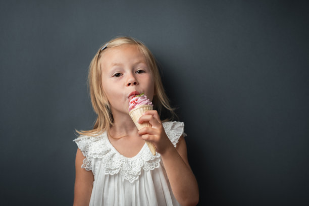 漂亮的白人小女孩，金发碧眼，穿着白色连衣裙，享受着灰色背景上的浆果味华夫筒冰淇淋图片下载