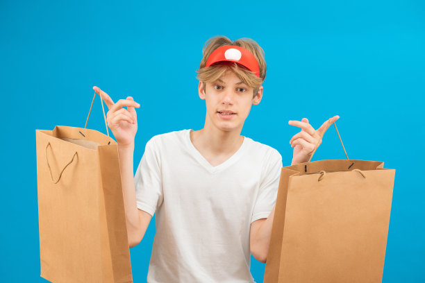 年轻小伙子在休闲白色t恤与纸袋孤立在蓝色背景的工作室肖像。人们生活方式的概念。模拟拷贝空间。图片下载