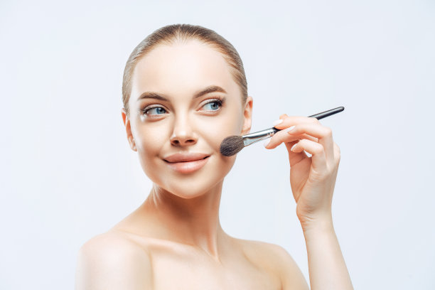 面部护理，美容理念。漂亮的年轻女子梳着深色的头发，用美容刷在脸上涂粉底，侧身站立，有裸露的身体，孤立在白色的背景上图片下载