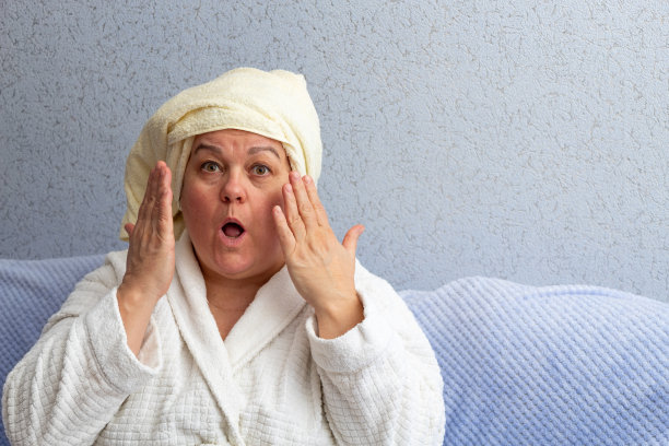 惊讶的老妇人与毛巾在她的头上和浴袍，她的手在她的脸和她的嘴是开放的。有选择性的重点。图片为与年龄相关的面部护理的文章。图片下载