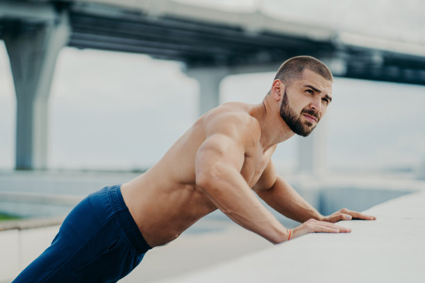户外拍摄的大胡子肌肉男做俯卧撑，在训练前热身，在桥的背景下摆姿势，在清晨锻炼，在某处集中精力。积极的生活方式图片下载