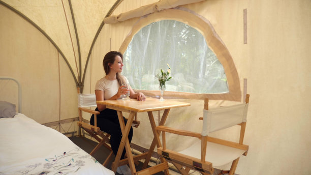 现代露营营内部。女人坐在豪华帐篷的窗边，喝着水，欣赏着大自然的景色。假期在营地。在森林中休息图片下载