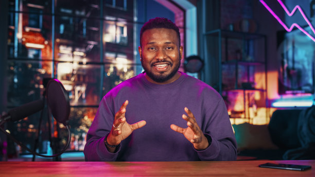 英俊的年轻非洲裔美国人对着麦克风说话，而录音广播节目从他的阁楼公寓。快乐的黑人男性在摄像机和录制播客上社交媒体直播。图片下载