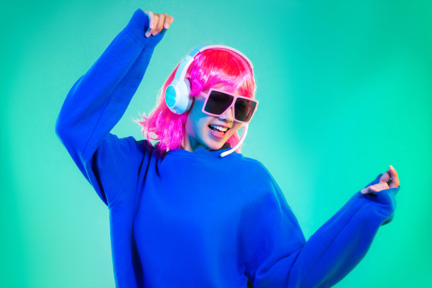 年轻的亚洲女子在蓝色运动衫粉红色短发朋克风格戴着墨镜和耳机在绿色背景上跳舞图片下载