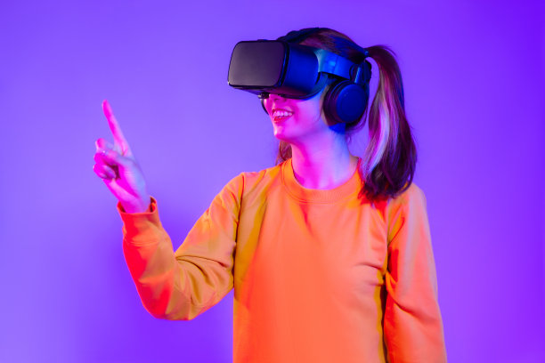 Metaverse的概念。年轻的亚洲女性穿着橙色运动衫，戴着黑色vr头盔观看触摸播放和手指指向紫色屏幕背景。图片下载