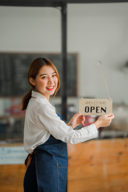 穿着围裙的美丽的亚洲咖啡师站在咖啡馆门口，门口的牌子是开着的。创业者的创业想法图片下载