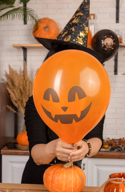 一个不知名的女人拿着一张可怕的万圣节脸在她的面前，一个亮橙色的气球。一个戴着狂欢节尖顶帽子的女人把脸藏在南瓜灯后面。图片下载