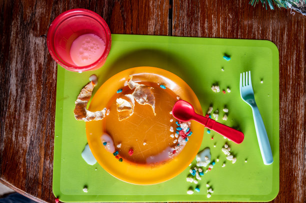 蹒跚学步的孩子吃完后，厨房桌子上留下的烂摊子。塑料叉子、杯子和盘子上撒满了水滴和面包屑。图片下载