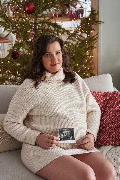 一个漂亮的孕妇穿着舒适的毛衣，坐在沙发上，旁边是一棵装饰着彩灯的漂亮圣诞树，手里拿着一张超声波照片。等待孩子的降生。怀孕的肚子图片下载